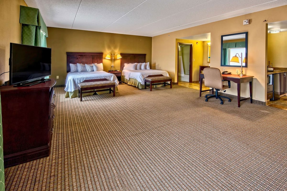 Quarto de hotel com duas camas, uma escrivaninha, uma cadeira, uma televisão e um banheiro.