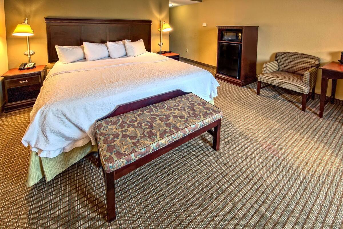 Quarto de hotel com uma cama grande, luminárias laterais, um banco de madeira, uma televisão e uma cadeira.