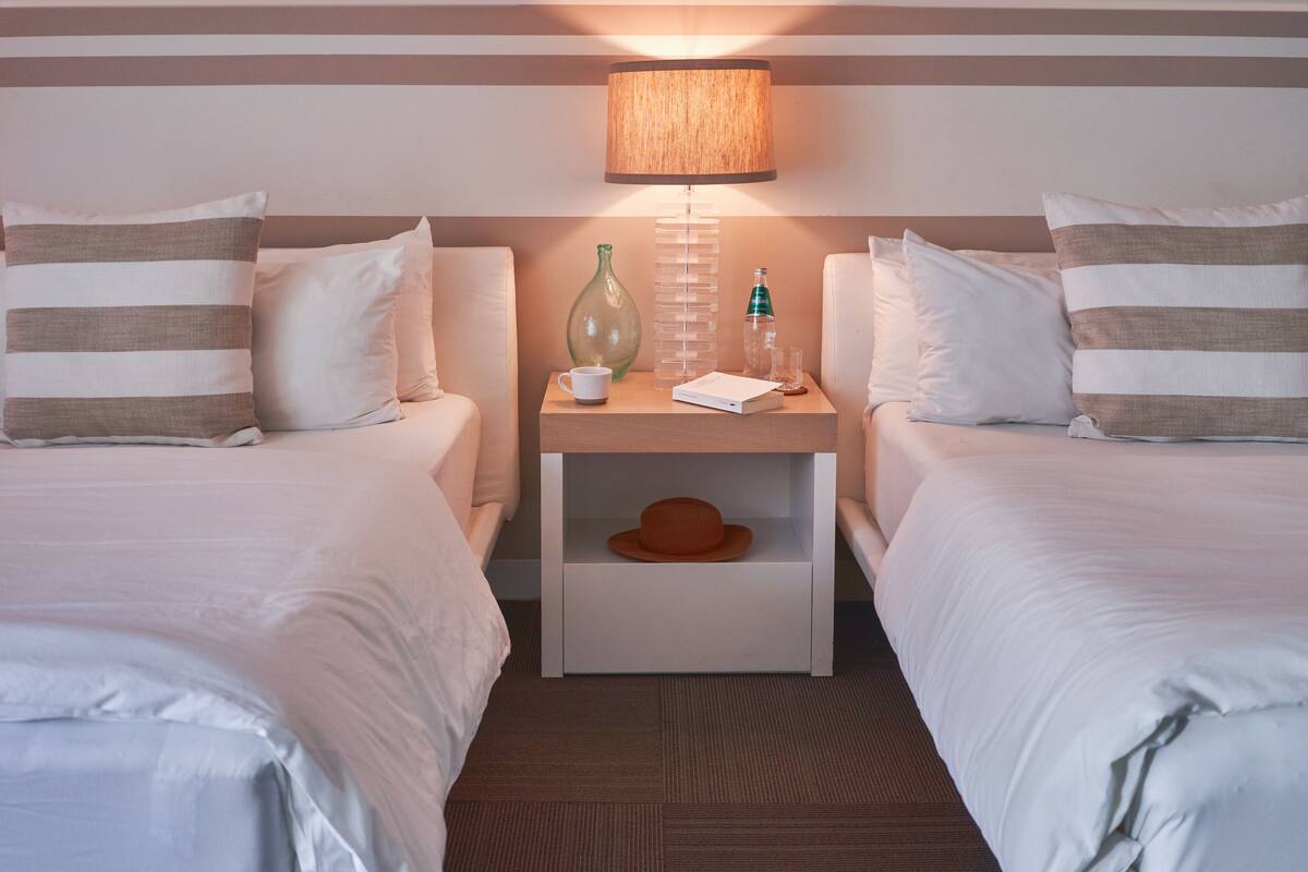 Une chambre d’hôtel avec deux lits, une table de chevet avec une lampe, un vase, une bouteille et un chapeau.