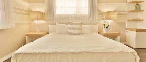 Une chambre d’hôtel bien aménagée, avec un grand lit, des lampes de chevet, une fenêtre ornée de rideaux légers et une petite étagère sur laquelle sont disposés des objets décoratifs.