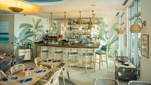 A bar area with wicker furniture, a bar counter with bottles, and a mural of tropical leaves. - The Local House (Miami Beach)