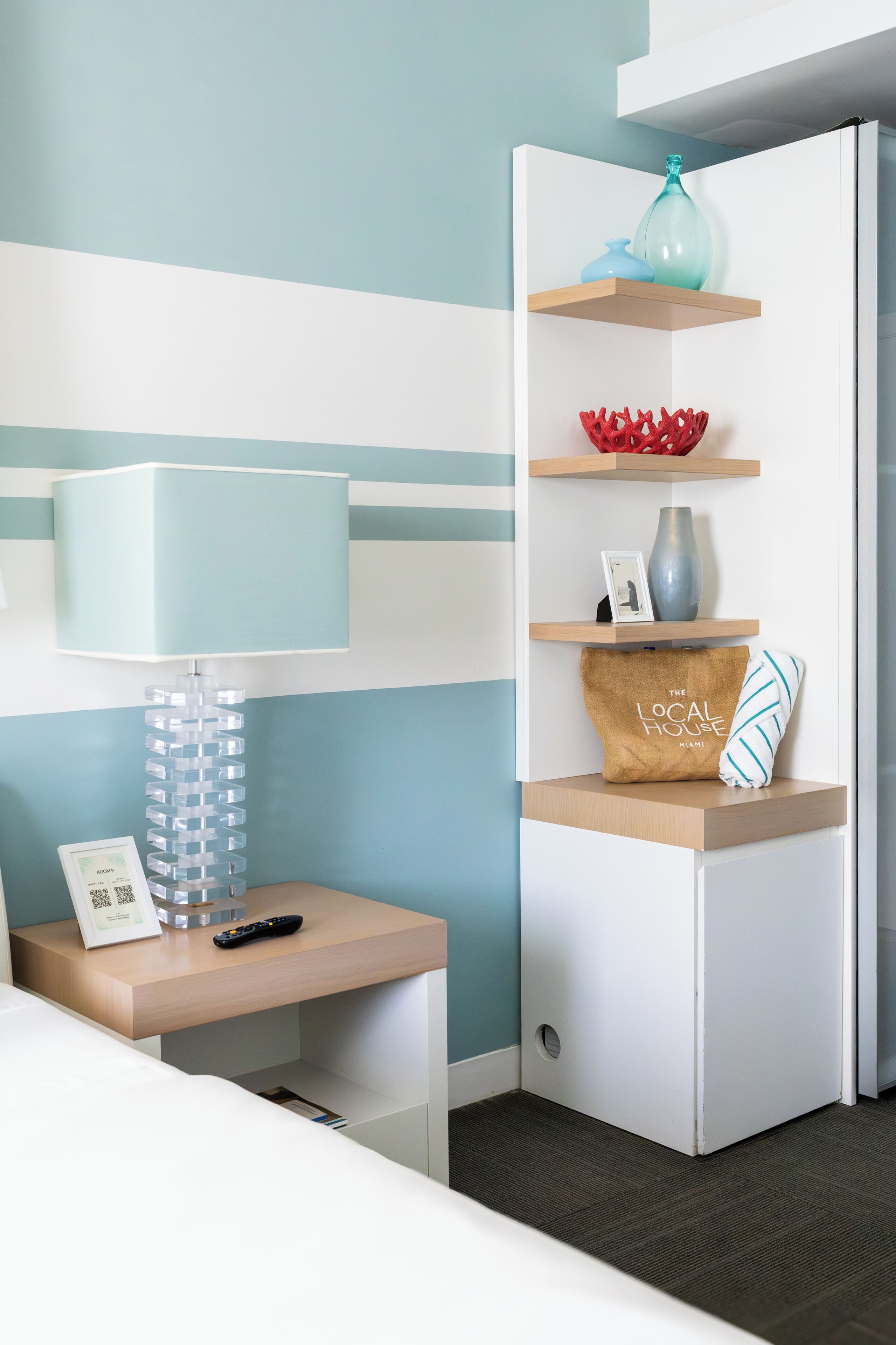 A hotel room with a bed, a bedside table with a lamp, a wall-mounted shelf with decorative items, and a door.