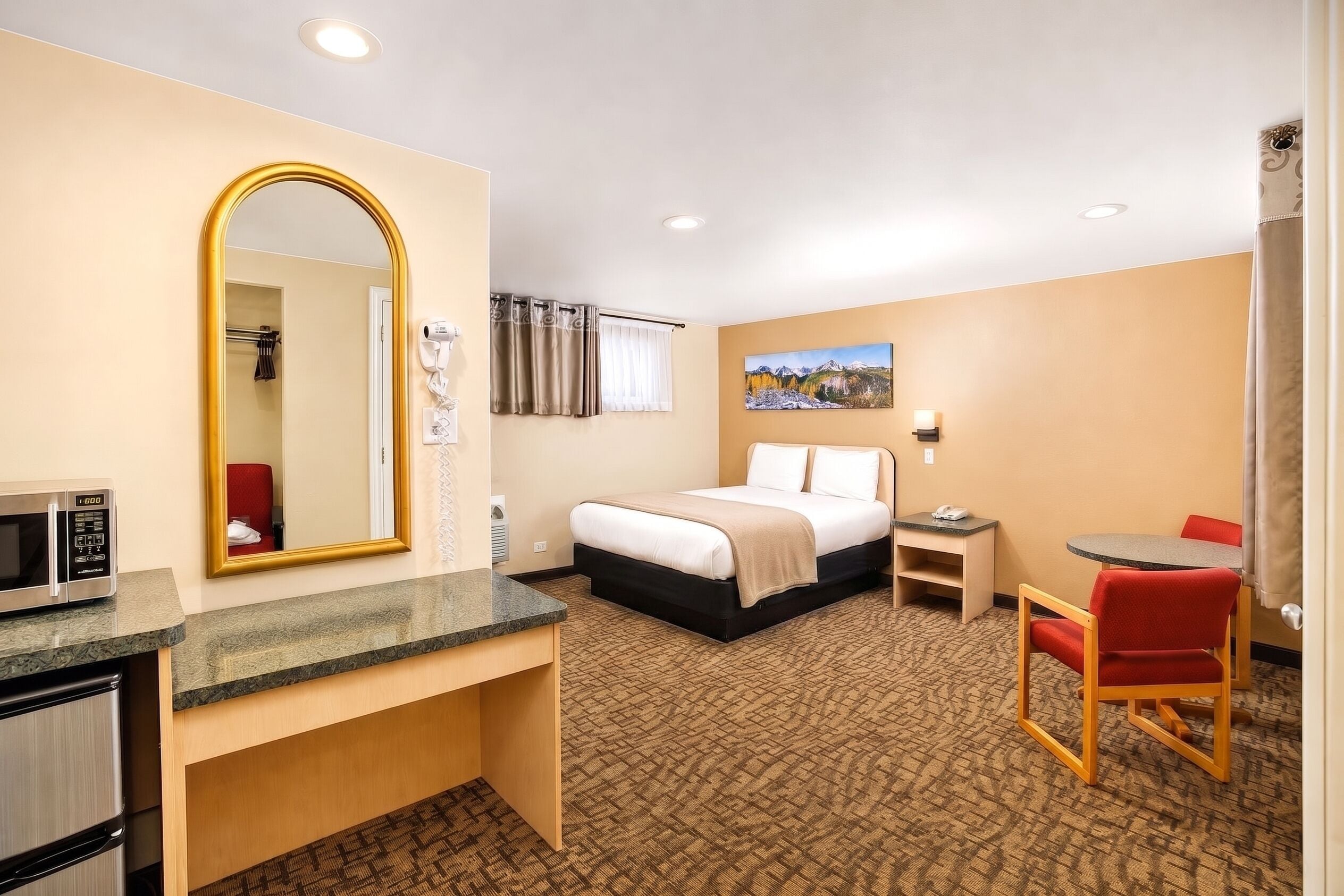 A hotel room with a bed, a desk with a microwave, a chair, a small table, and a painting on the wall.