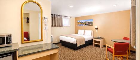 A hotel room with a bed, a desk with a microwave, a chair, a small table, and a painting on the wall.