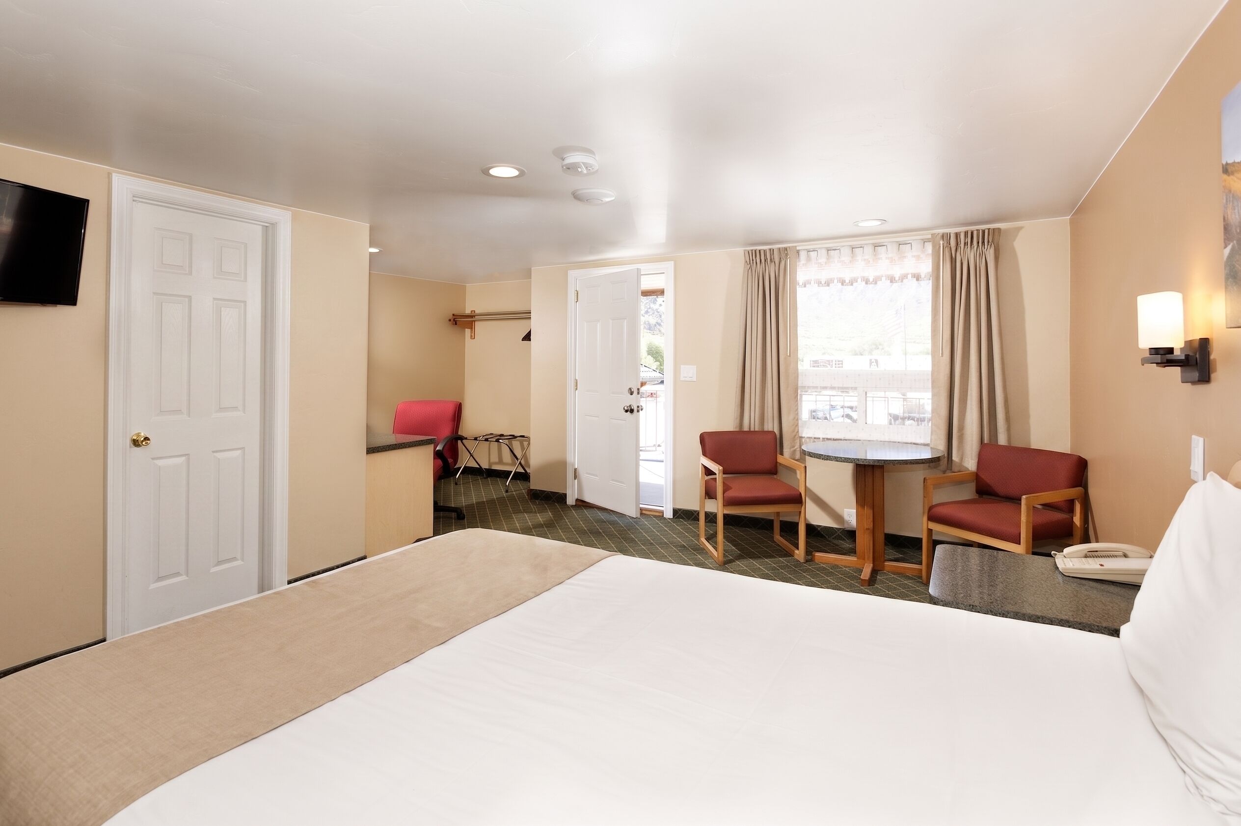 A hotel room with a bed, two red chairs, a small table, a TV, and a window with curtains.