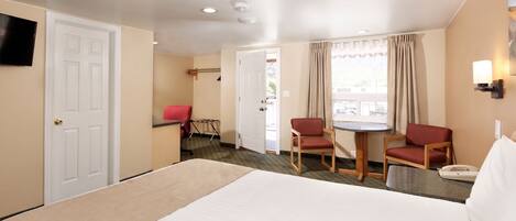 A hotel room with a bed, two red chairs, a small table, a TV, and a window with curtains.