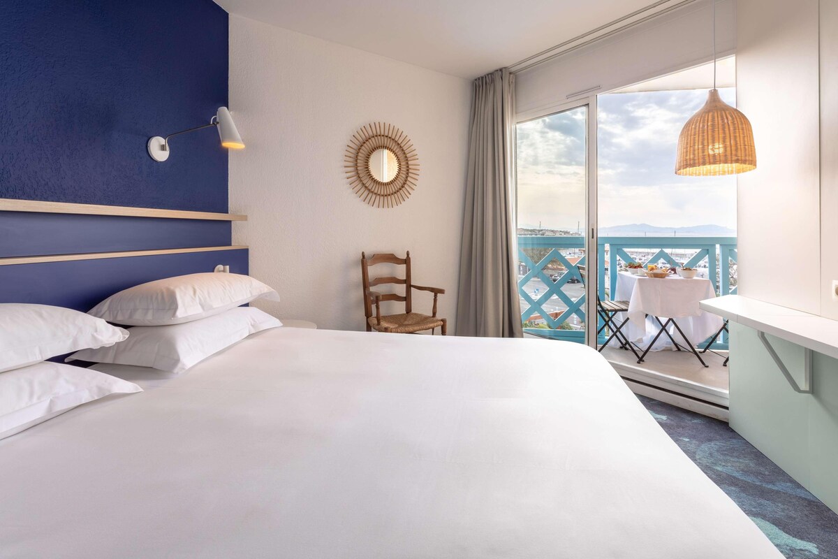 A hotel room with a large bed, a chair, a table with a view, and a balcony with a railing.
