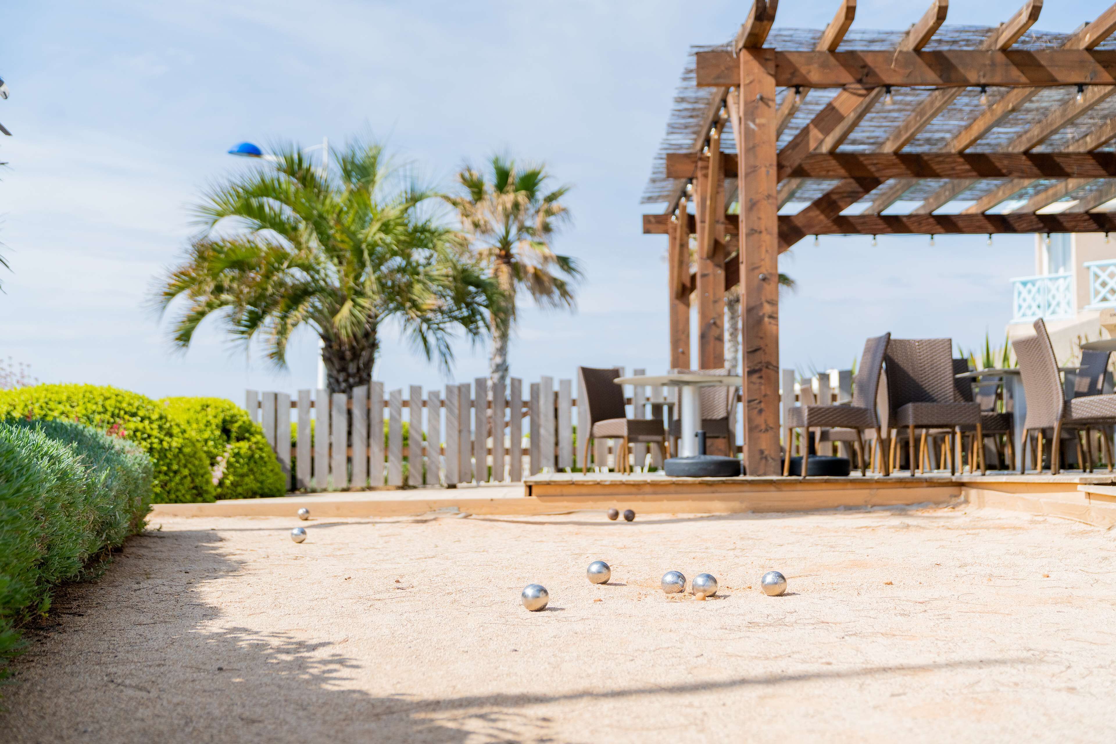 A sunny outdoor area with a wooden pergola, palm trees, and a seating area with chairs and tables.