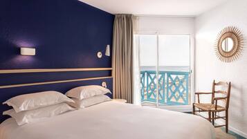 Una camera d'albergo con un letto grande, una sedia di legno, vista sul mare e un balcone con ringhiera blu.