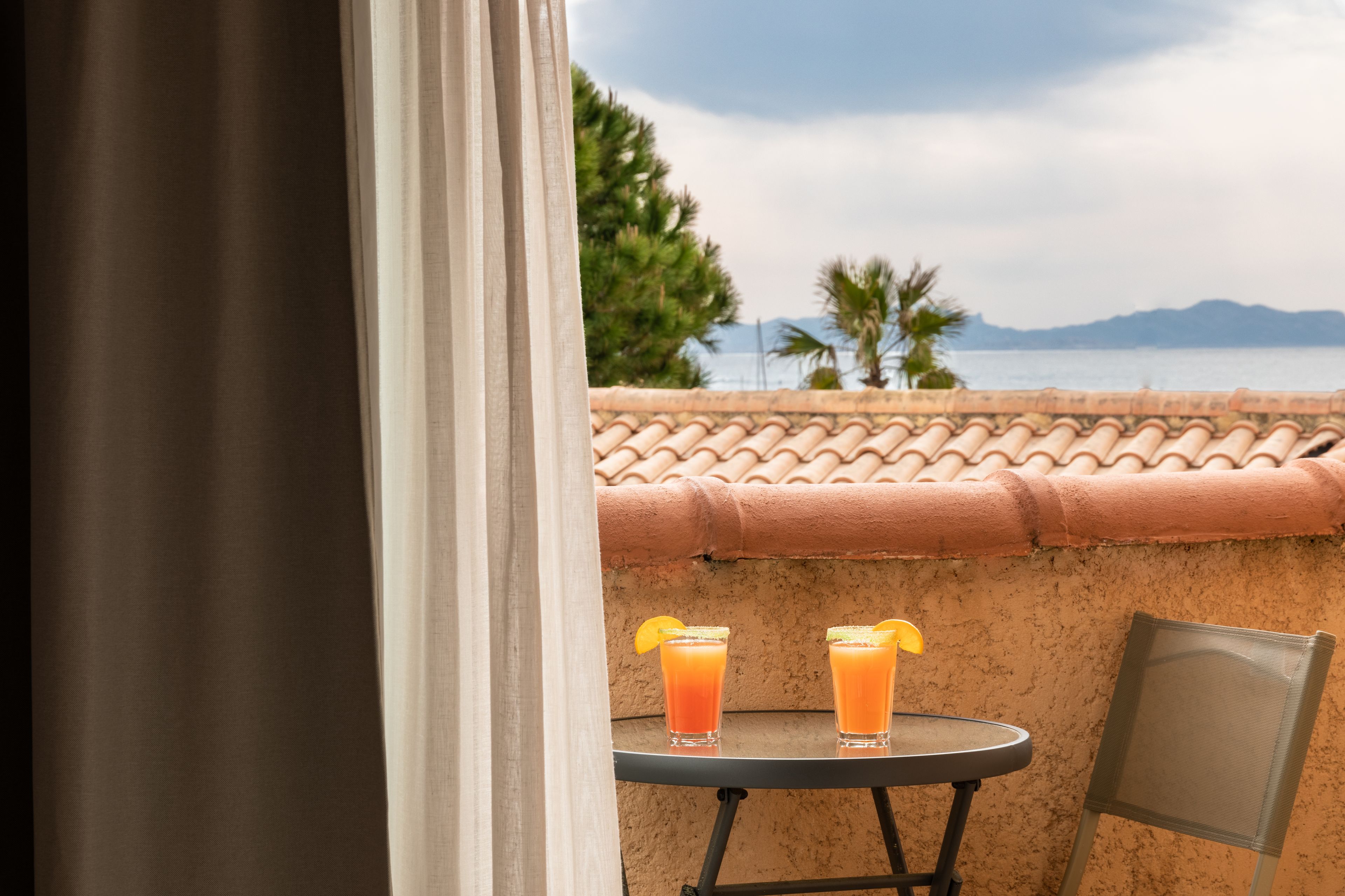 Un balcón con una mesa y dos vasos de jugo de naranja, vista al mar y una montaña al fondo.