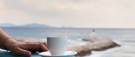 A white cup on a saucer on a blue table by the sea with a rocky outcrop in the background.