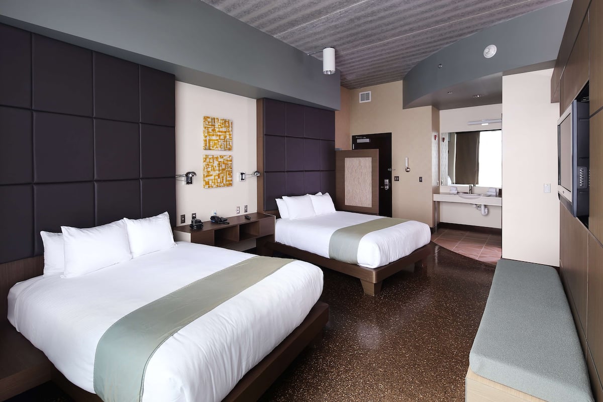 Een hotelkamer met twee bedden, een grote spiegel, een bankje en een badkamer met een wastafel en toilet.