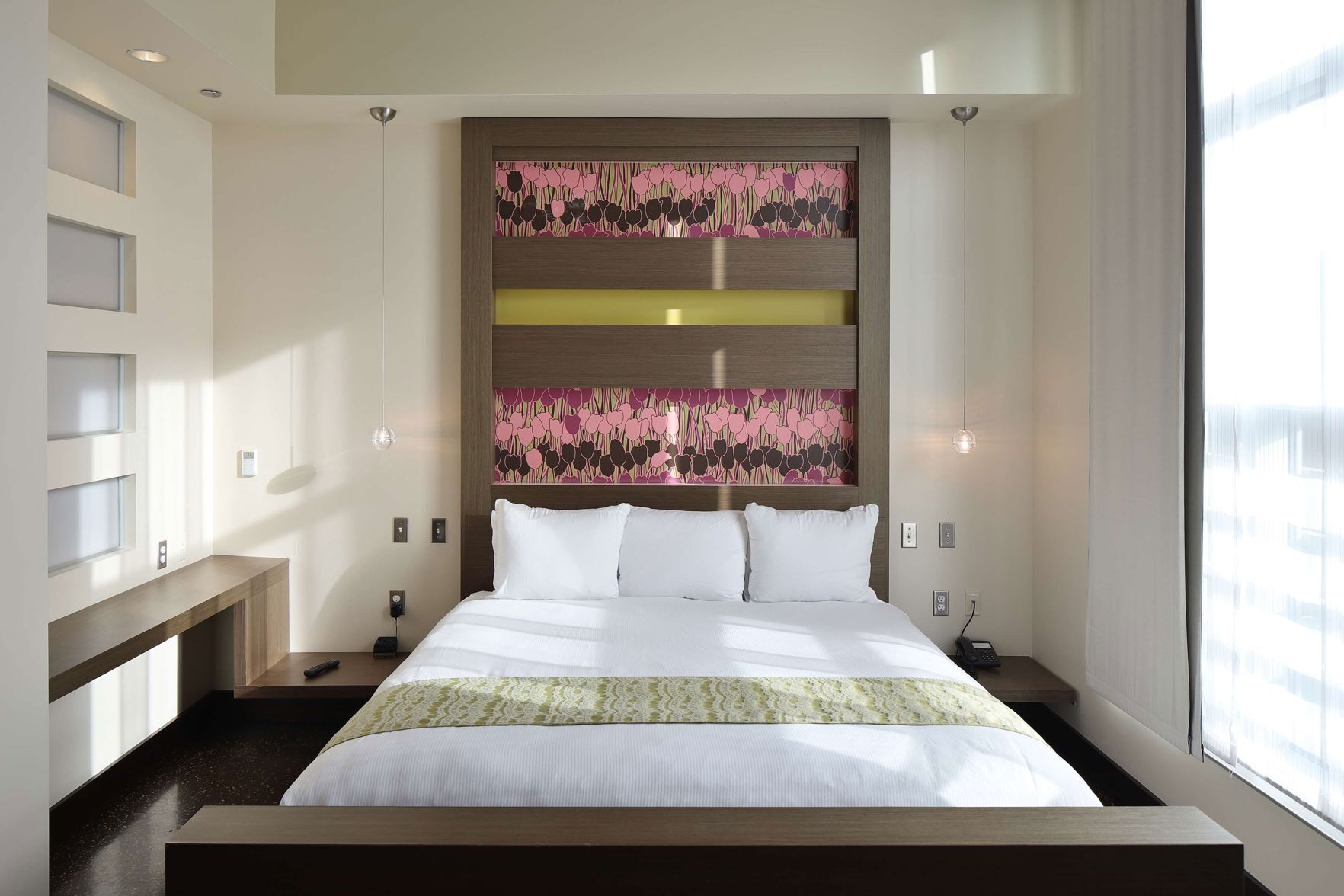 Een moderne hotelkamer met een groot bed, een hoofdbord met bloemenprint, inbouwkasten, nachtkastjes en een groot raam.