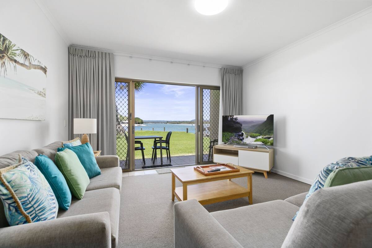 A modern living room with a sofa, coffee table, and TV. There is a sliding glass door leading to a balcony with a view of the sea.
