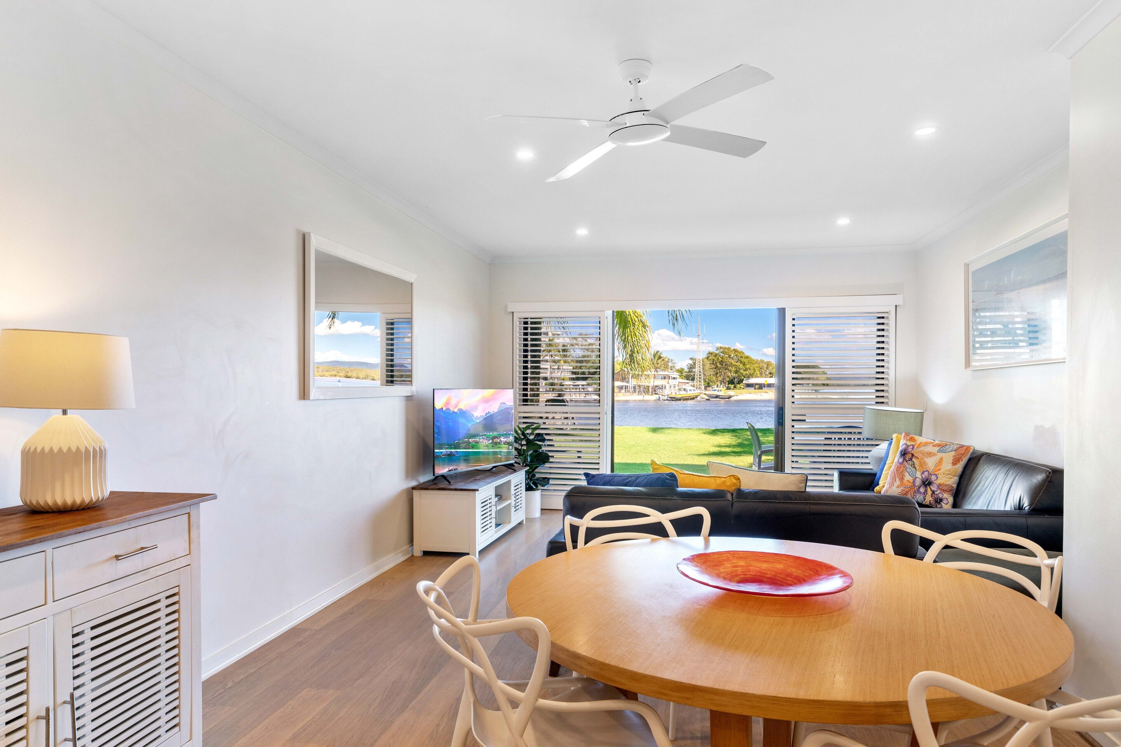 A modern living room with a dining table, white chairs, a sofa, and a ceiling fan.