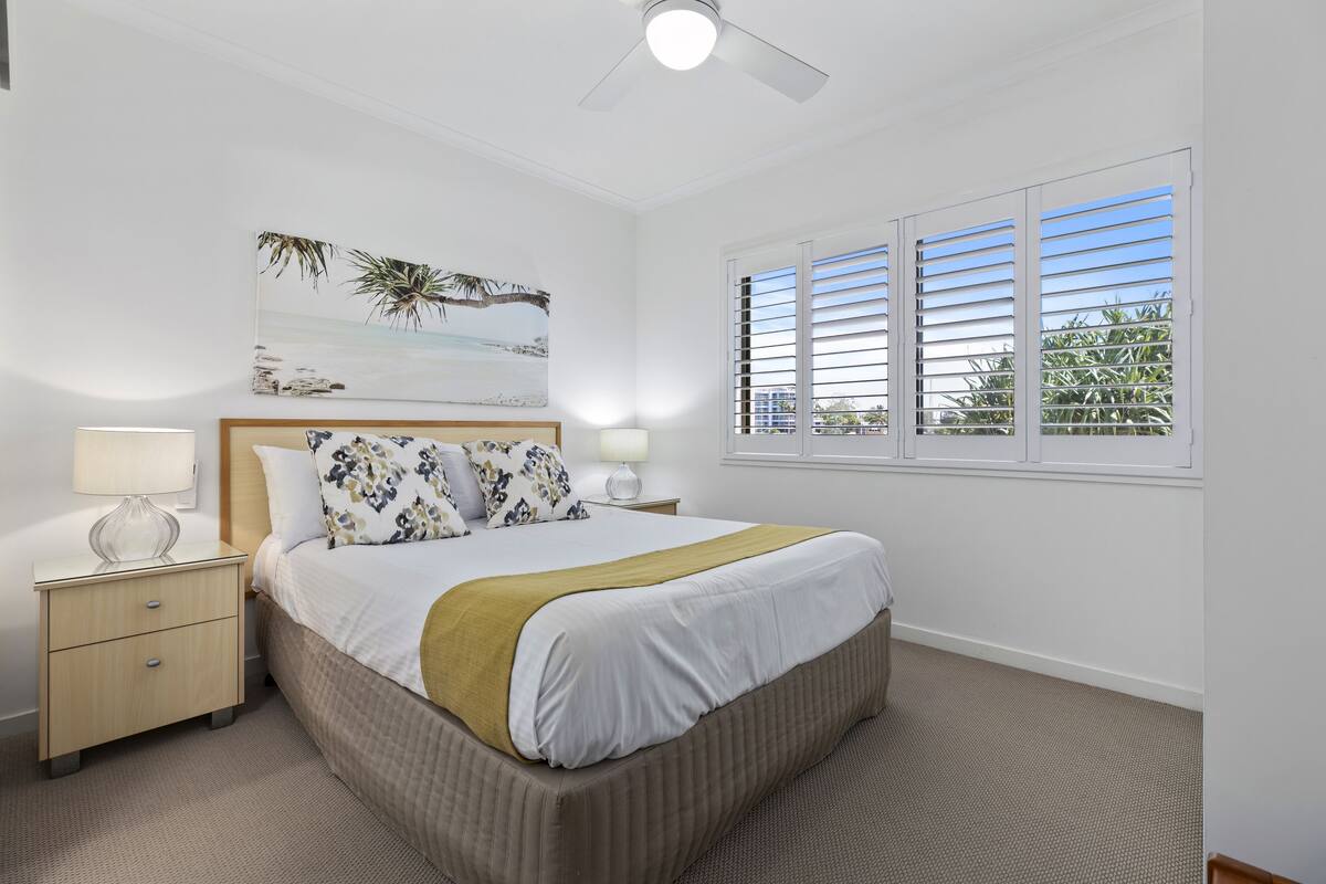 A bedroom with a bed, a nightstand, a lamp, a ceiling fan, a window with blinds, and a beach-themed painting on the wall.