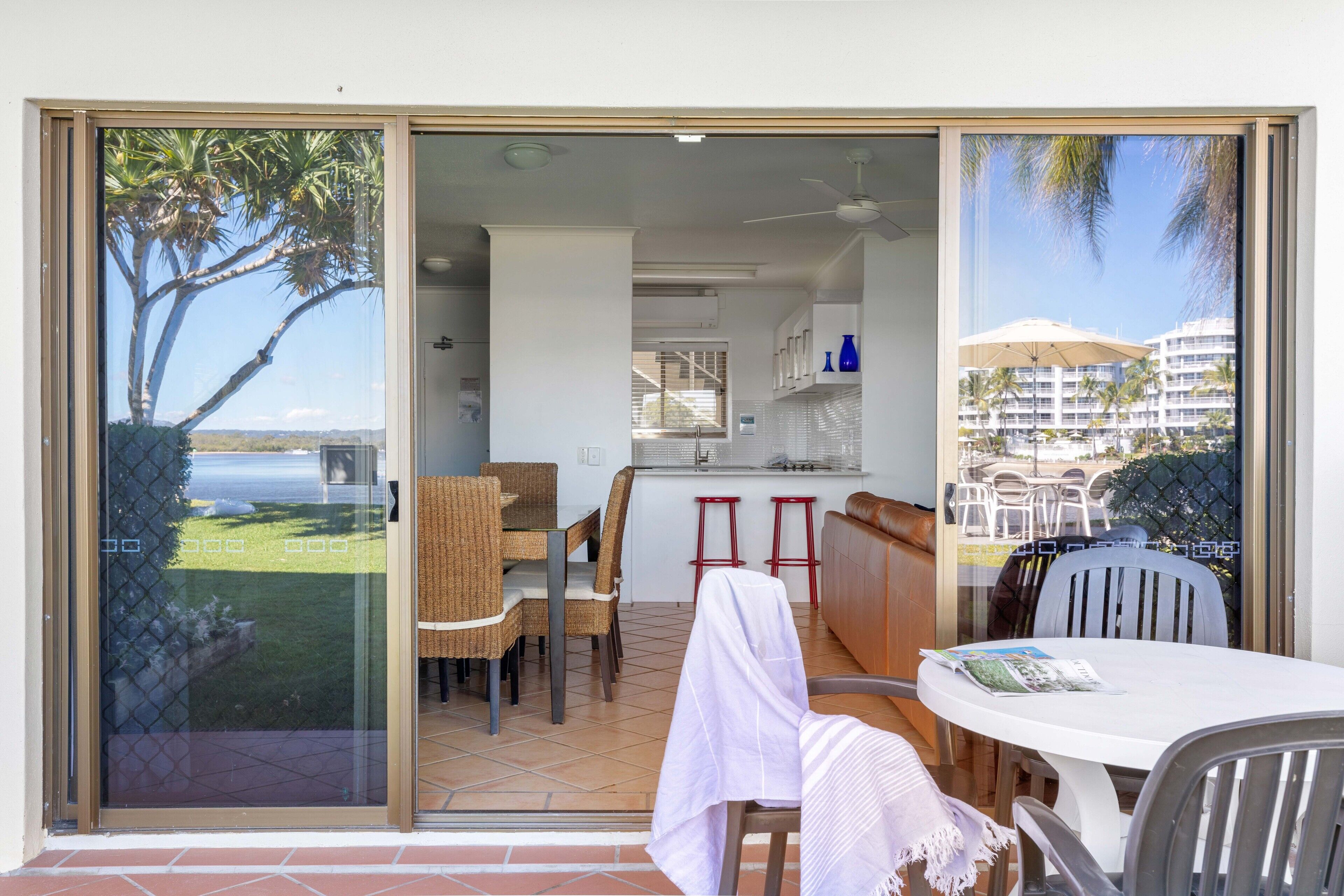 A dining area with a table and chairs, a kitchen area with a counter and stools, and a view of palm trees and a body of water through a glass door.