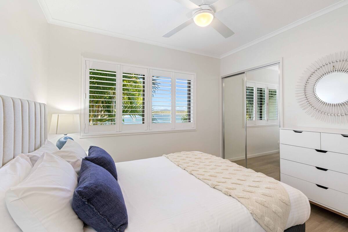A bedroom with a bed, a ceiling fan, a window with blinds, a dresser, and a mirror.