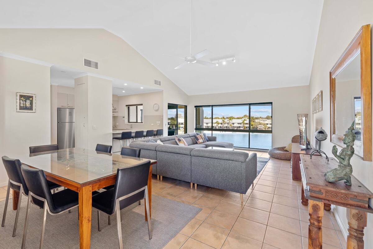 A spacious living area with a glass-top dining table, grey sectional sofa, and a large window offering a view of the sea.