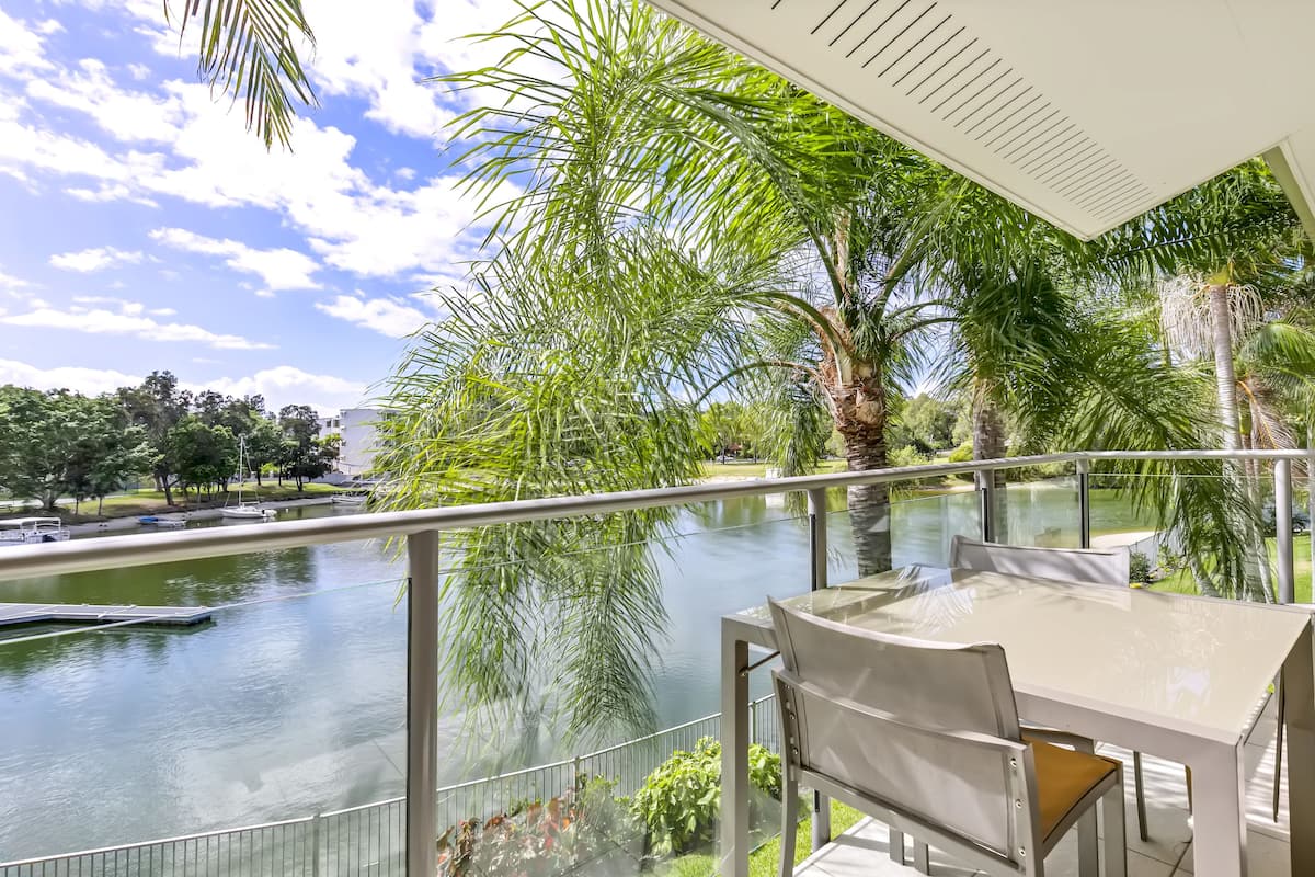 A balcony with a glass railing overlooking a body of water, surrounded by lush greenery and palm trees.