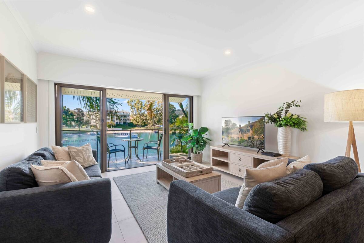 A modern living room with a sofa, coffee table, and TV. Large windows offer a view of a pool and outdoor seating.