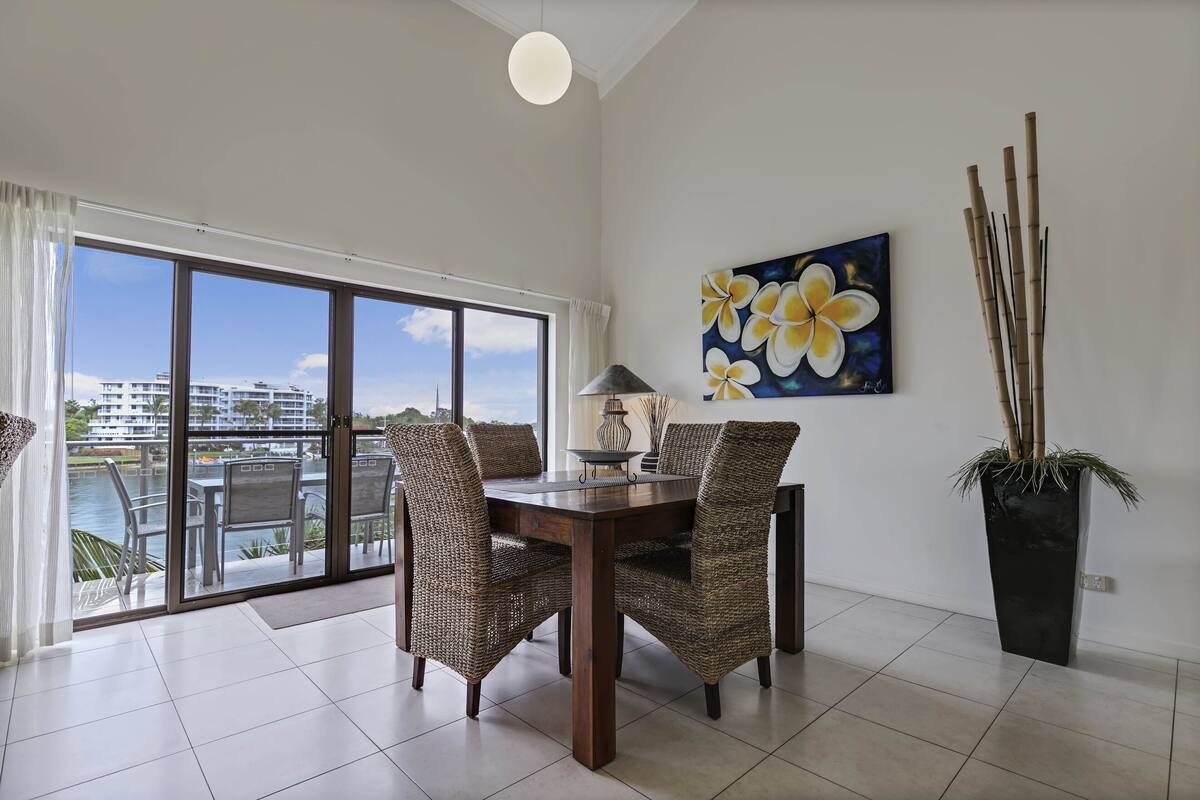A dining area with a view of a body of water and buildings, featuring wicker chairs, a table, and a potted plant.