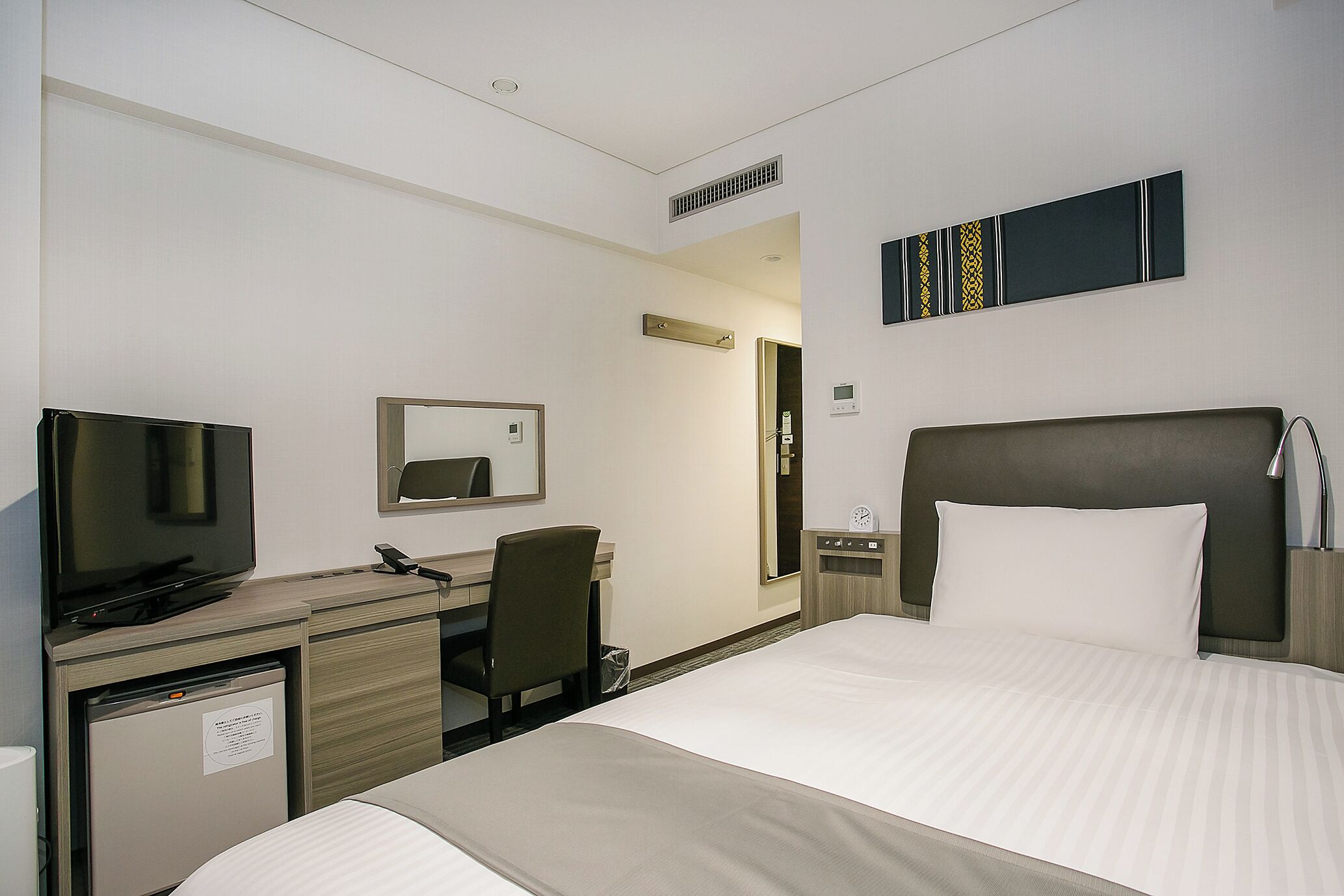 A hotel room with a bed, a desk with a television, a chair, a mirror, a nightstand, and a wall-mounted clock.