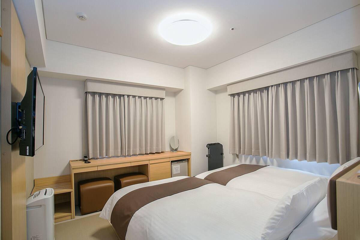 Ein Hotelzimmer mit zwei Betten, einem Schreibtisch, einem Stuhl, einem Fernseher und einem Fenster mit Vorhängen.