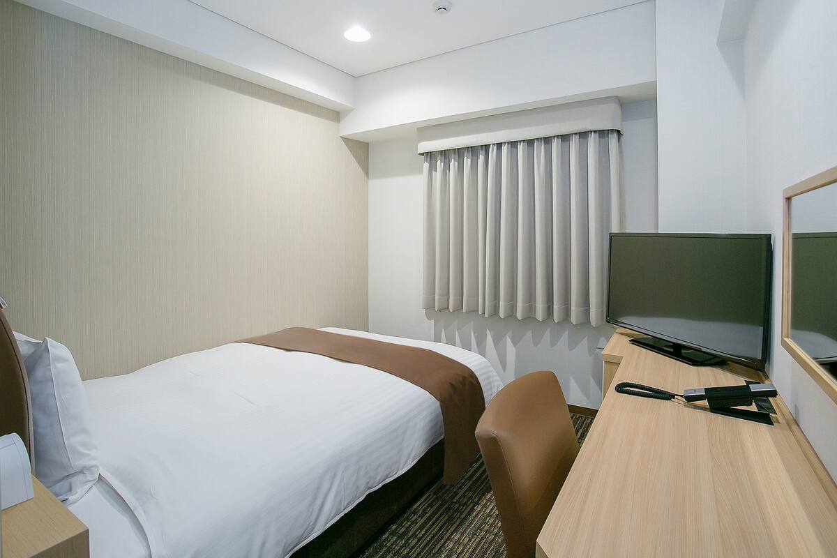 Ein Hotelzimmer mit Bett, Schreibtisch, Stuhl, Fernseher und einem Fenster mit Vorhängen.