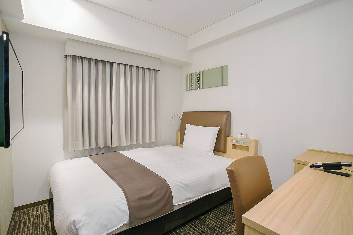 Ein Hotelzimmer mit Bett, Schreibtisch, Stuhl, einem Fenster mit Vorhängen und einem an der Wand befestigten Fernseher.