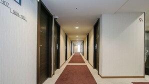 Ein langer, schmaler Hotelgang mit rotem Teppich, Türen an beiden Seiten und einem Zimmerschild an der linken Wand.