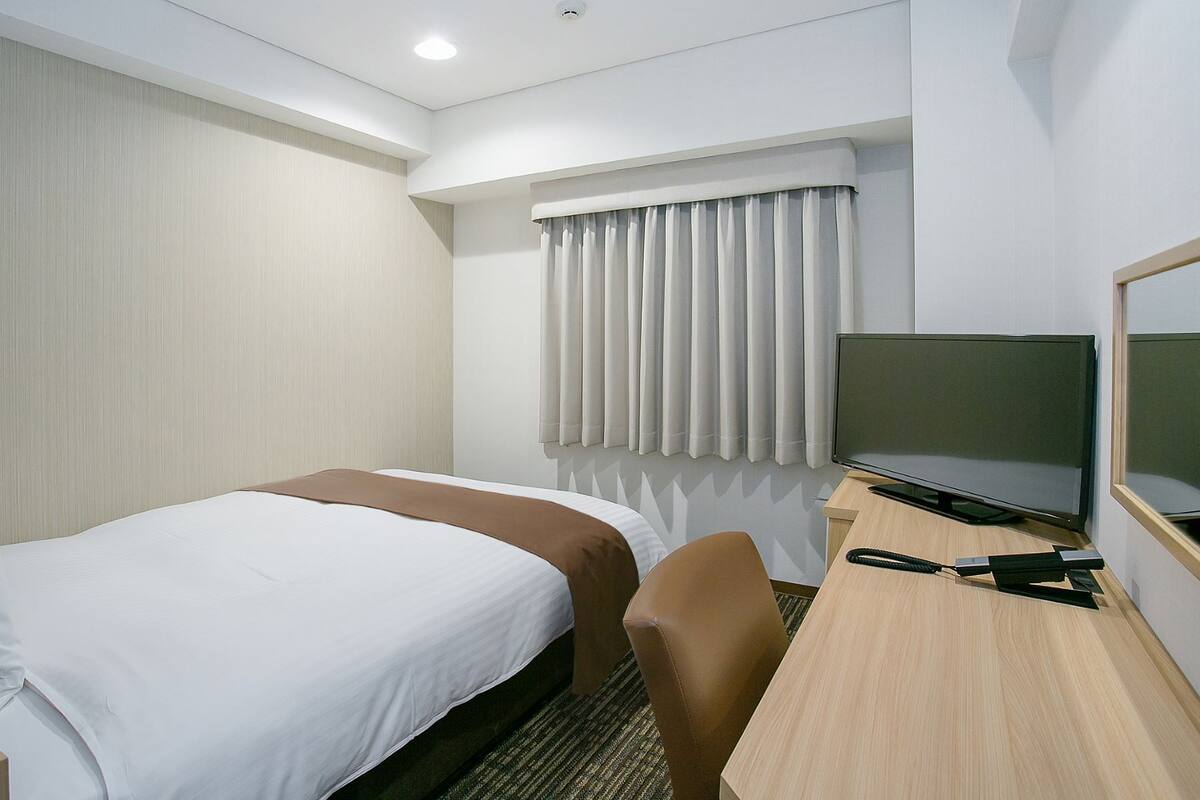 Ein Hotelzimmer mit einem Bett, einem Schreibtisch mit Fernseher, einem Sessel und einem Fenster mit Vorhängen.