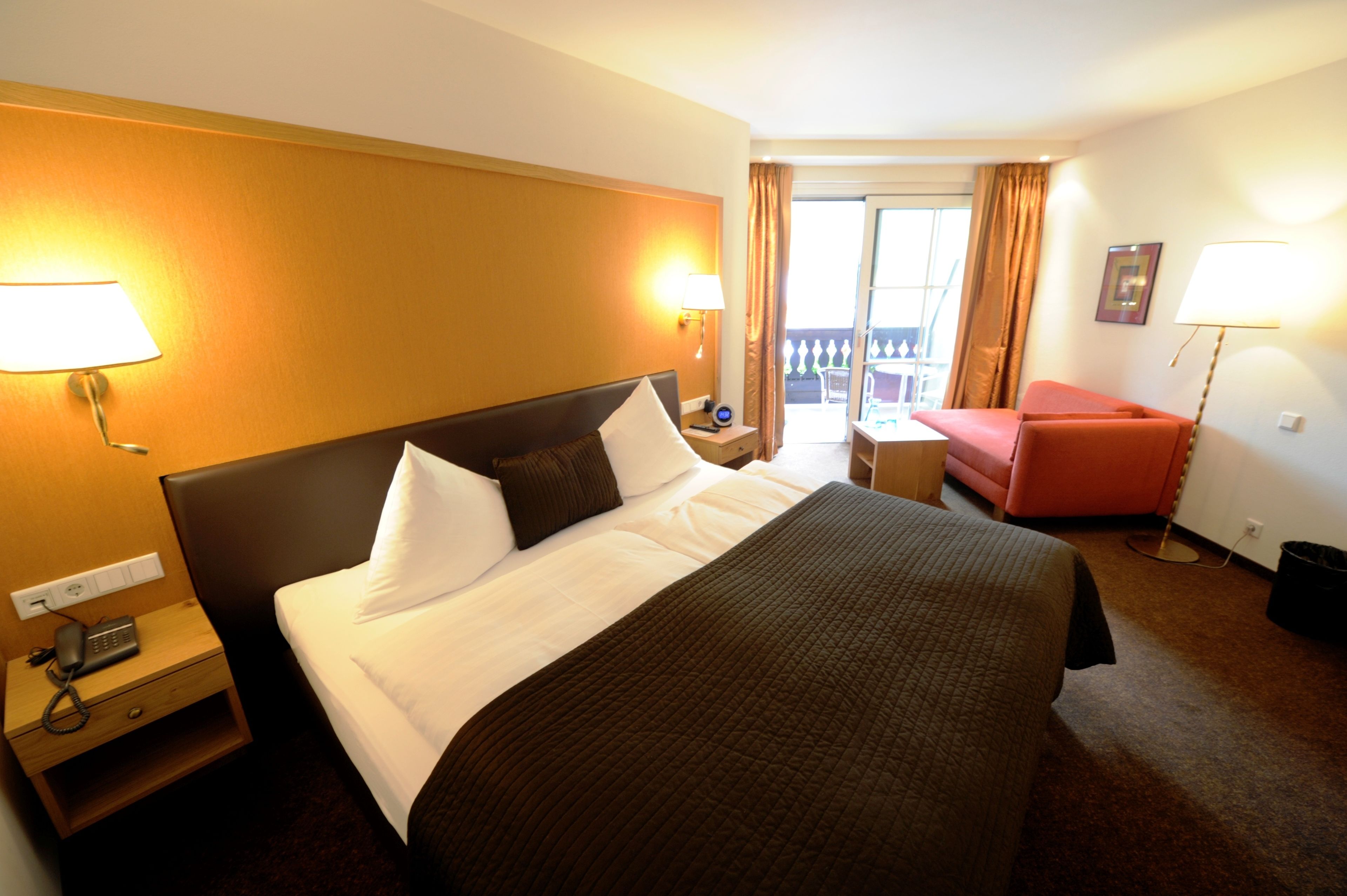 A hotel room with a large bed, a nightstand with a phone, a red chair, and a balcony with curtains.