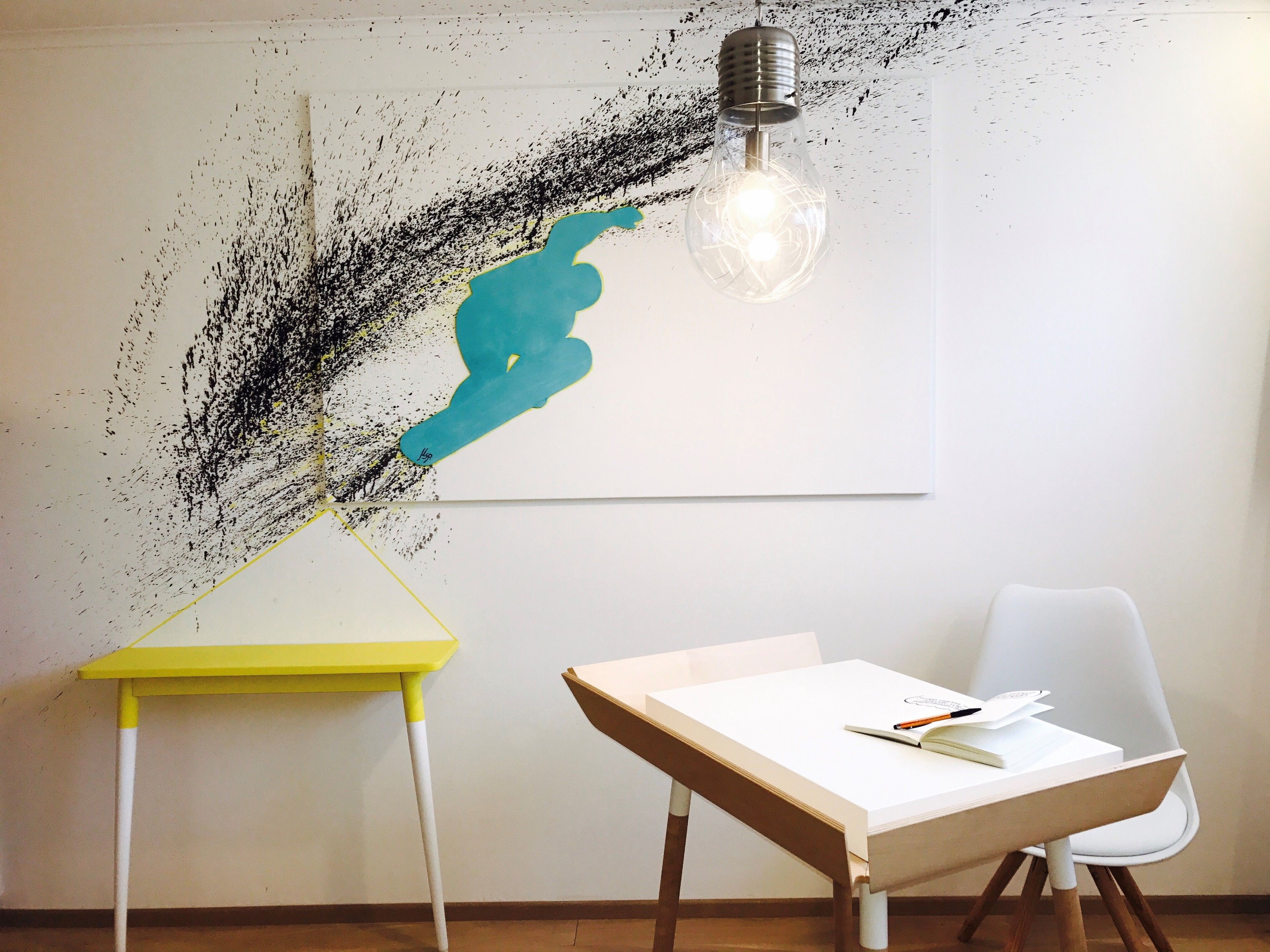 A room with a white table, a yellow table, and a white chair. There is a painting on the wall and a light fixture above the table.