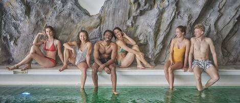 Un grupo de personas sentadas en el borde de una piscina, con un muro de roca al fondo.