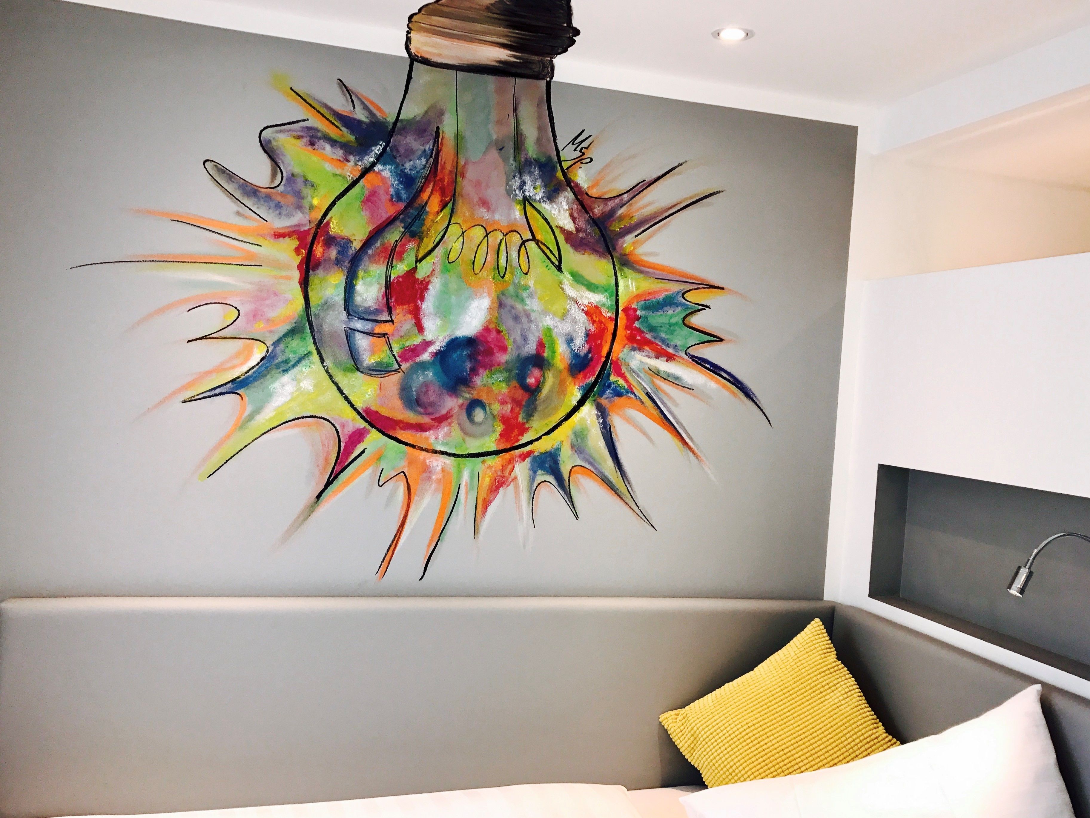 A modern living room with a colorful light bulb artwork on the wall, a grey sofa, and a yellow pillow.