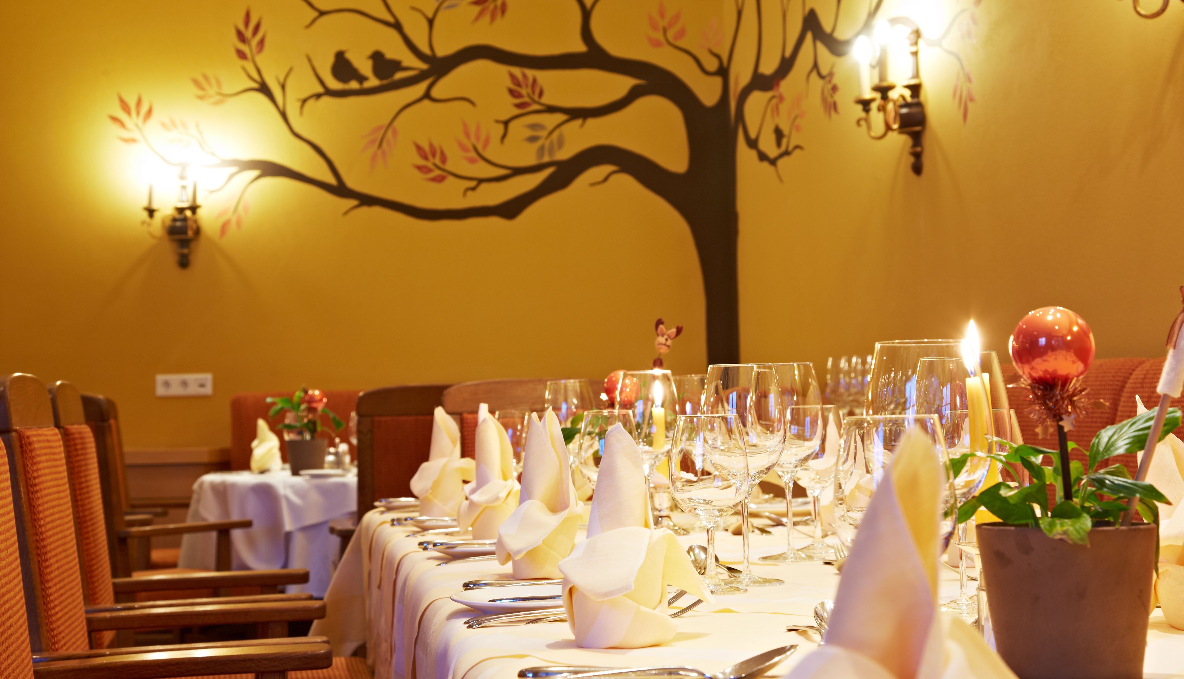 A restaurant setting with a long table set for dining, candle-lit wall sconces, and a tree mural on the wall.