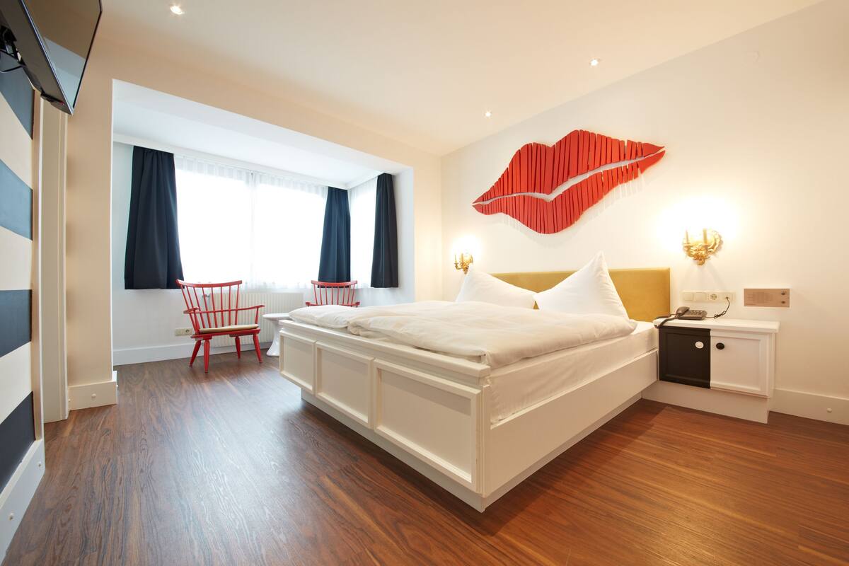 Una habitación de hotel moderna con una cama grande, una mesita de noche con lámpara, una silla roja y una ventana con cortinas.