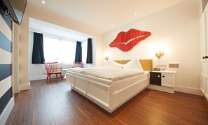 Une chambre d’hôtel moderne avec un grand lit, une table de chevet avec une lampe, un fauteuil rouge et une fenêtre avec des rideaux.