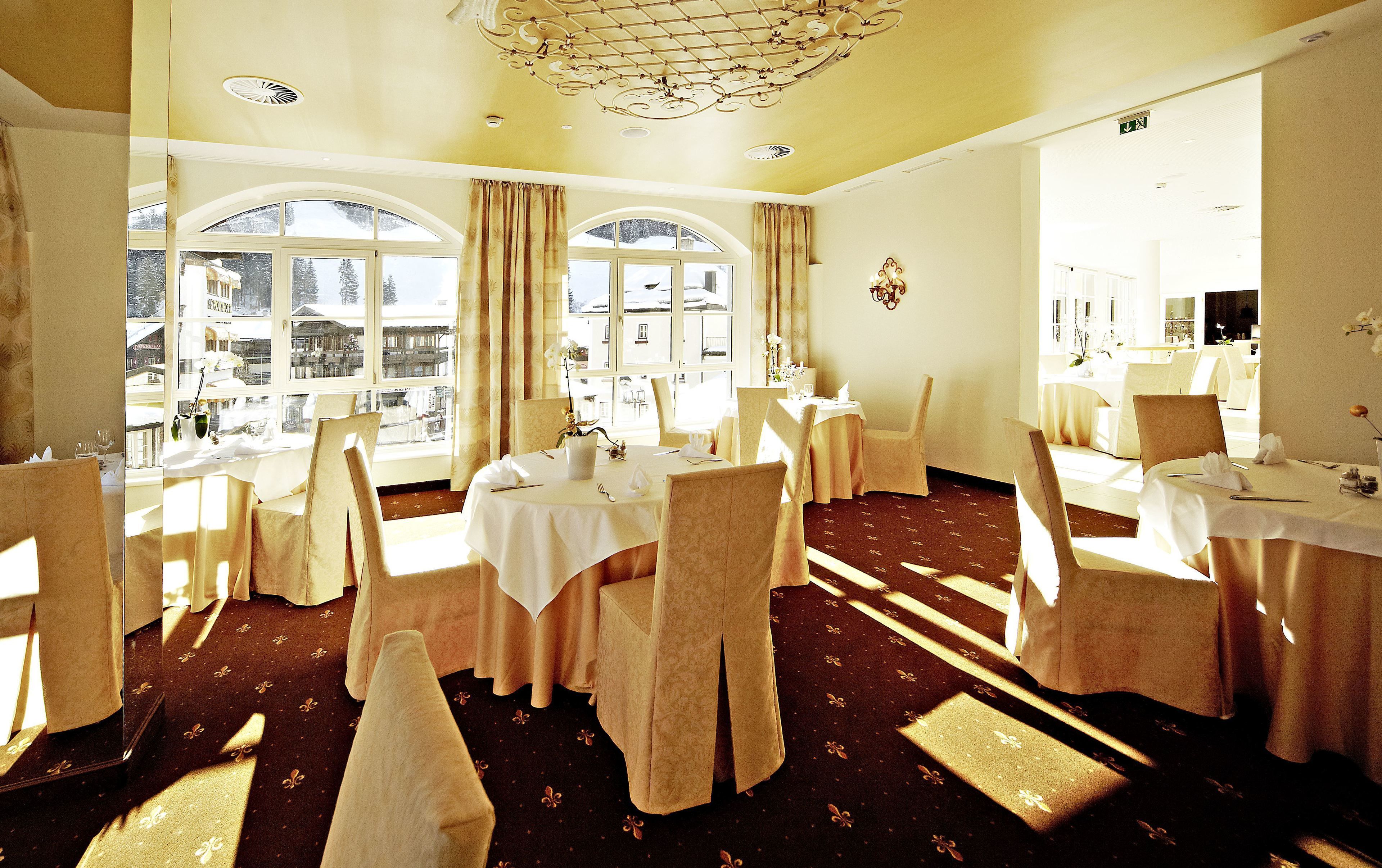 A hotel dining area with multiple tables and chairs, large windows with curtains, and a chandelier.