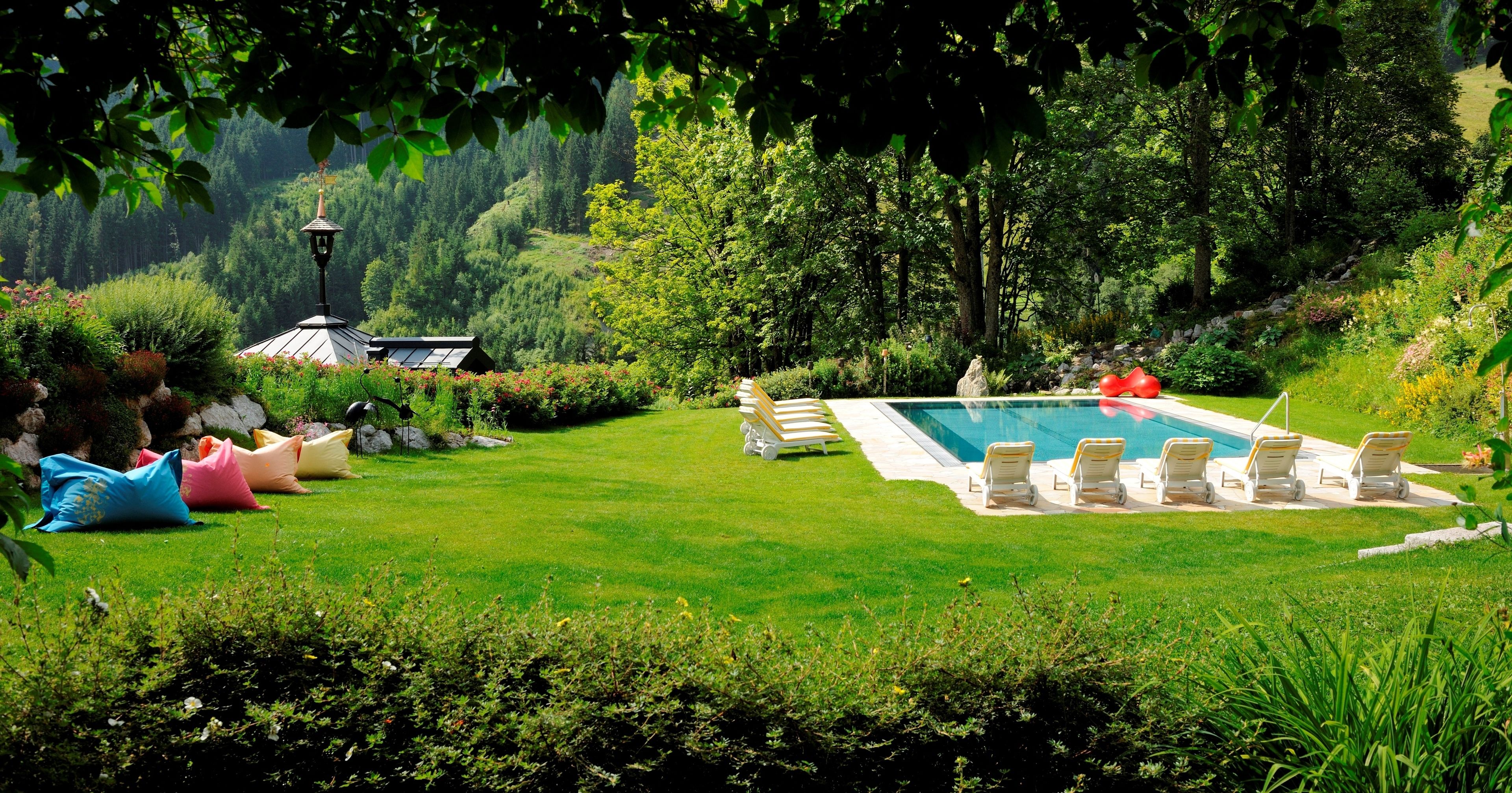 Uma área de piscina com espreguiçadeiras, um poste de luz e um gazebo num jardim exuberante.
