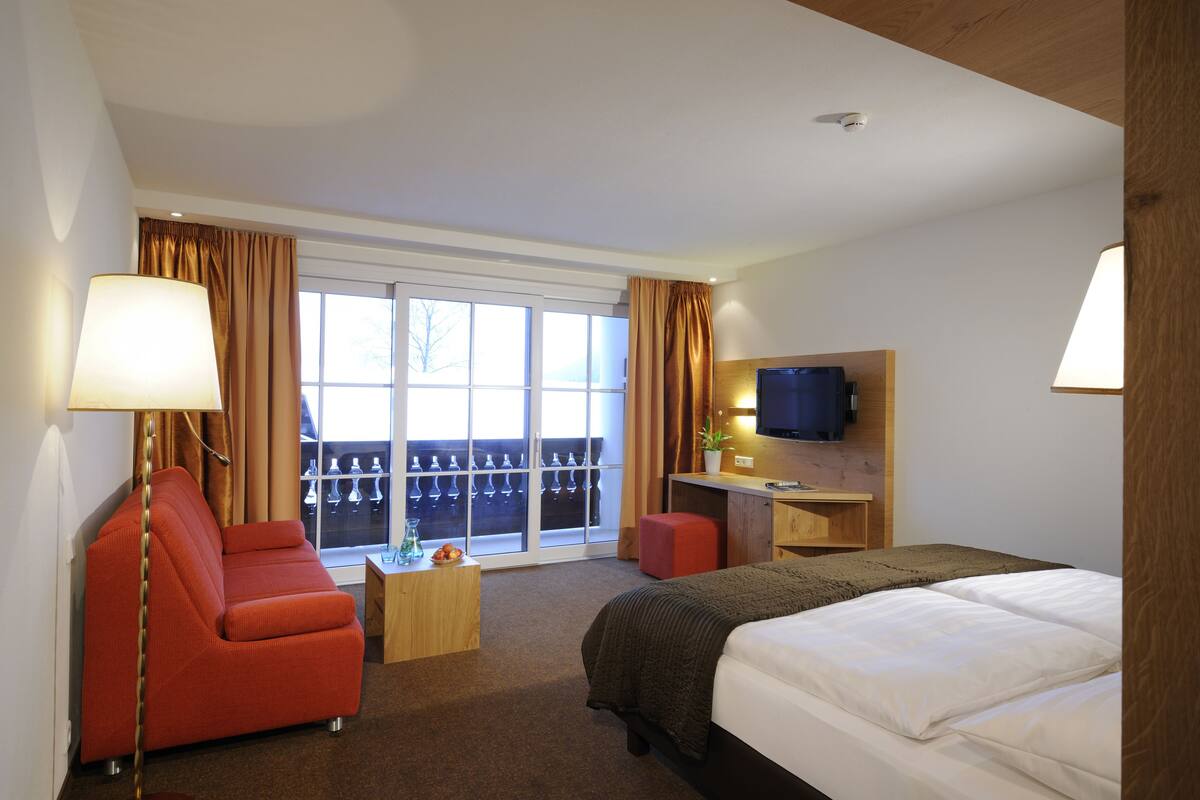 Habitación de hotel con cama, escritorio, televisor, sillón rojo y ventana con cortinas.