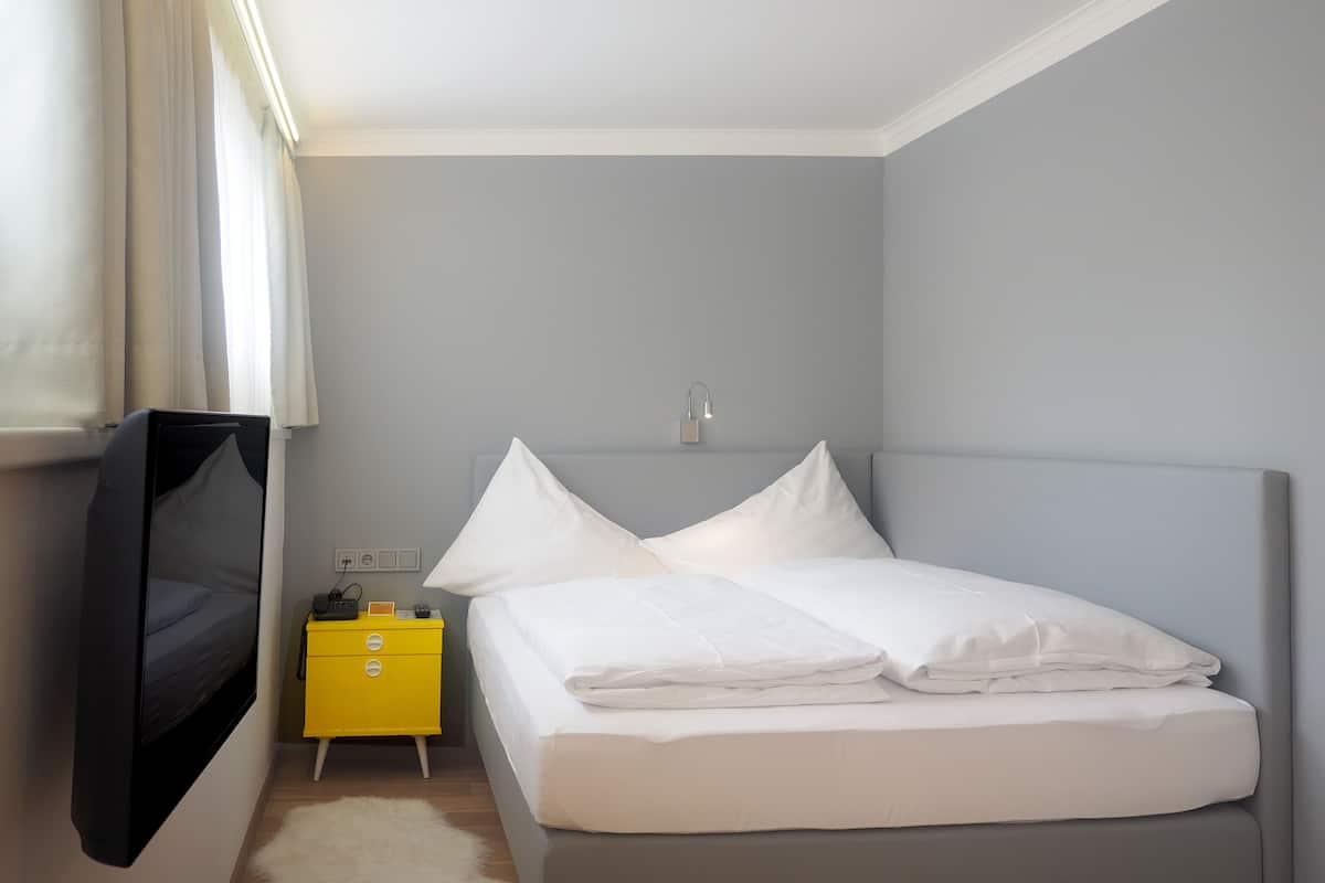 Un dormitorio con una cama, televisor montado en la pared, una mesita de noche y un tocador.