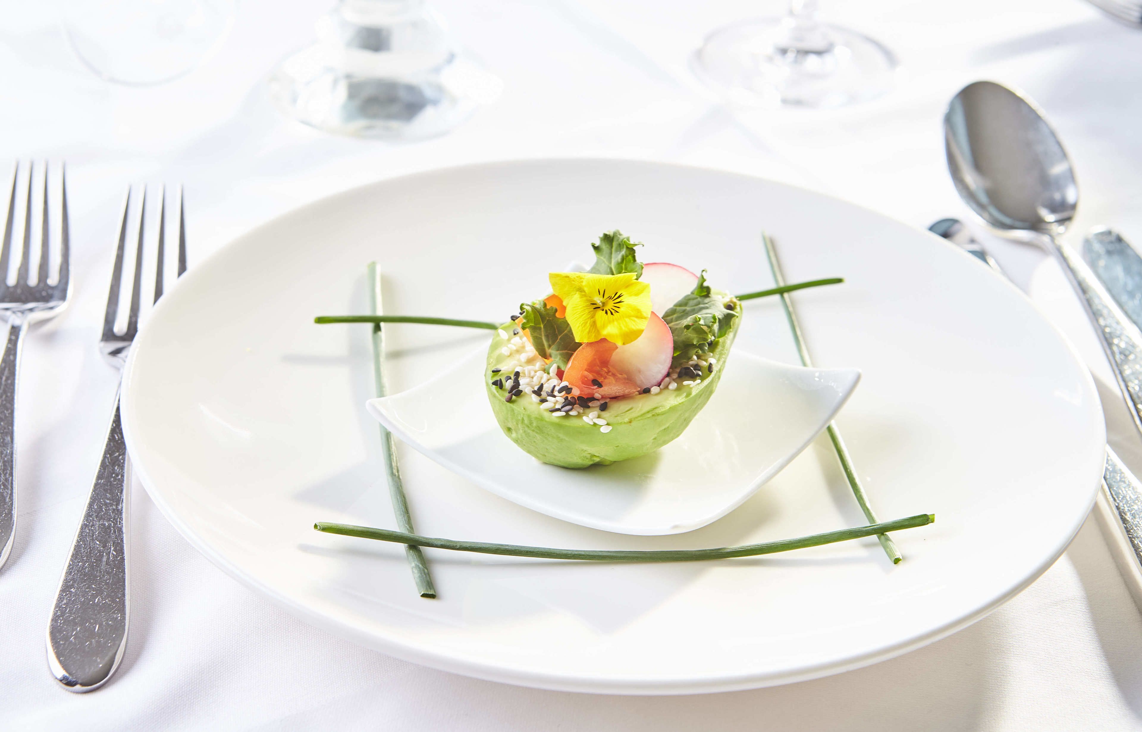 A white plate with a green vegetable-based dish, garnished with a flower and radish, on a white tablecloth.