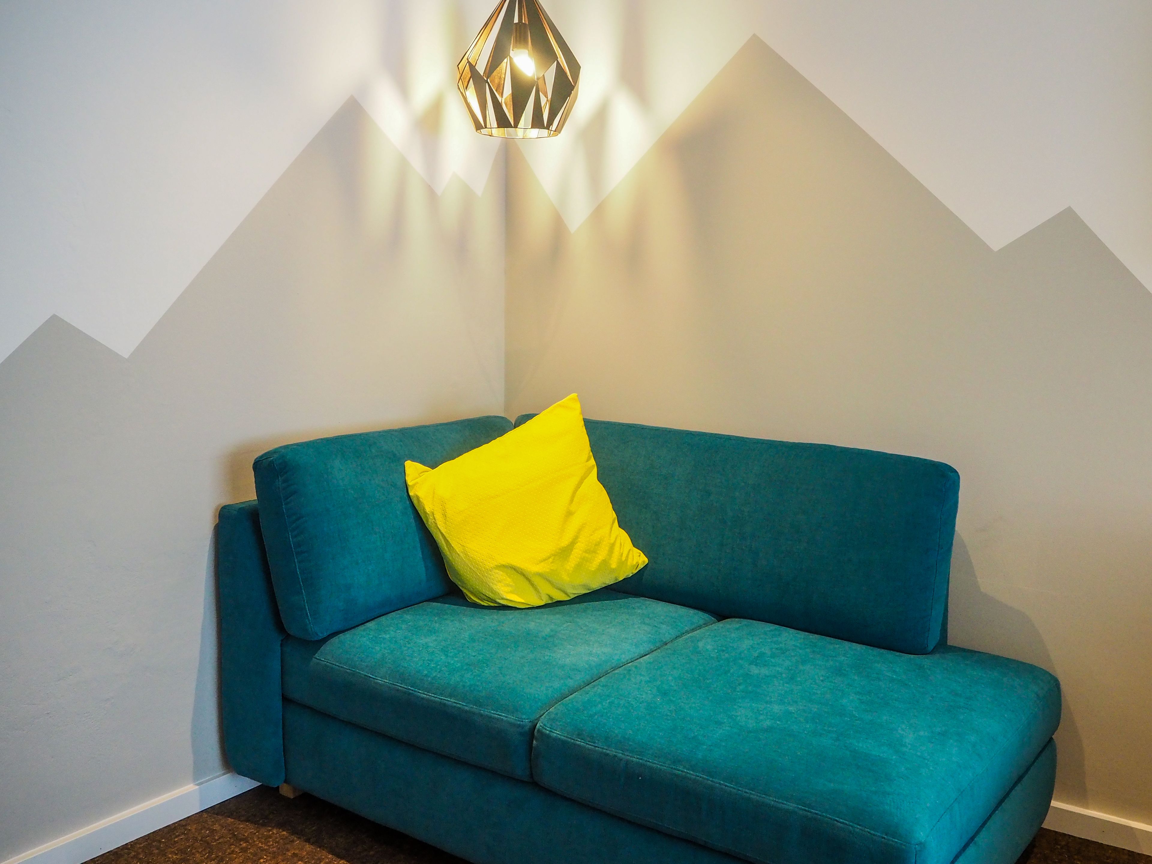 A teal sofa with a yellow pillow against a white wall with a geometric pattern.