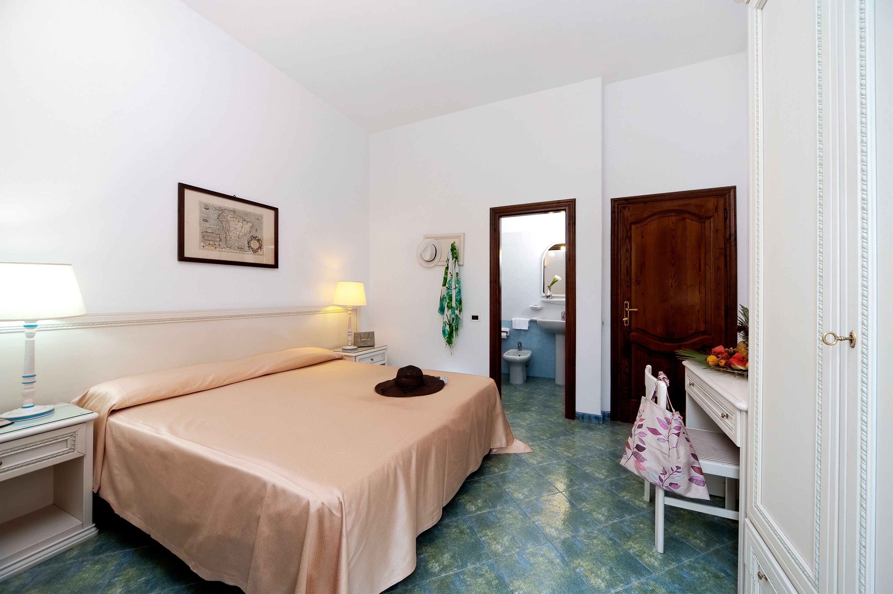 A hotel room with a bed, a nightstand, a lamp, a framed map on the wall, a mirror, a wooden door, and a bathroom visible through an open door.