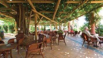 Un café al aire libre con mobiliario de madera y suelo de baldosas, rodeado de árboles.