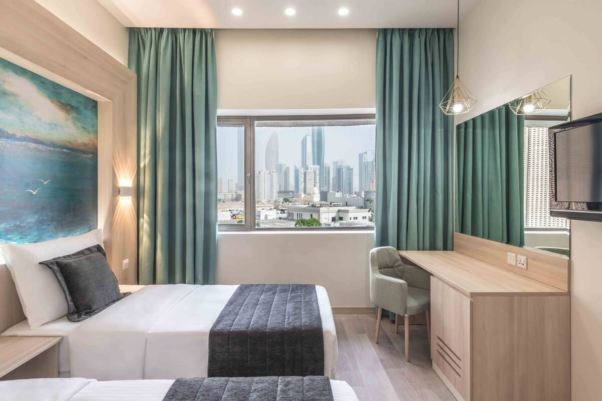 A hotel room with a bed, a desk, a chair, a large window with a city view, and a painting on the wall.