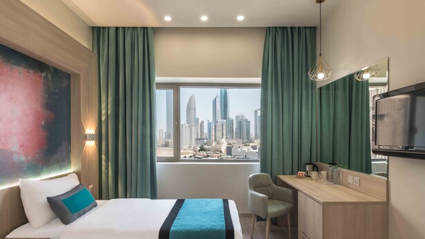 A hotel room with two beds, a desk, a chair, a TV, and a large window with a city view. - Oasis Hotel Kuwait (Kuwait City)