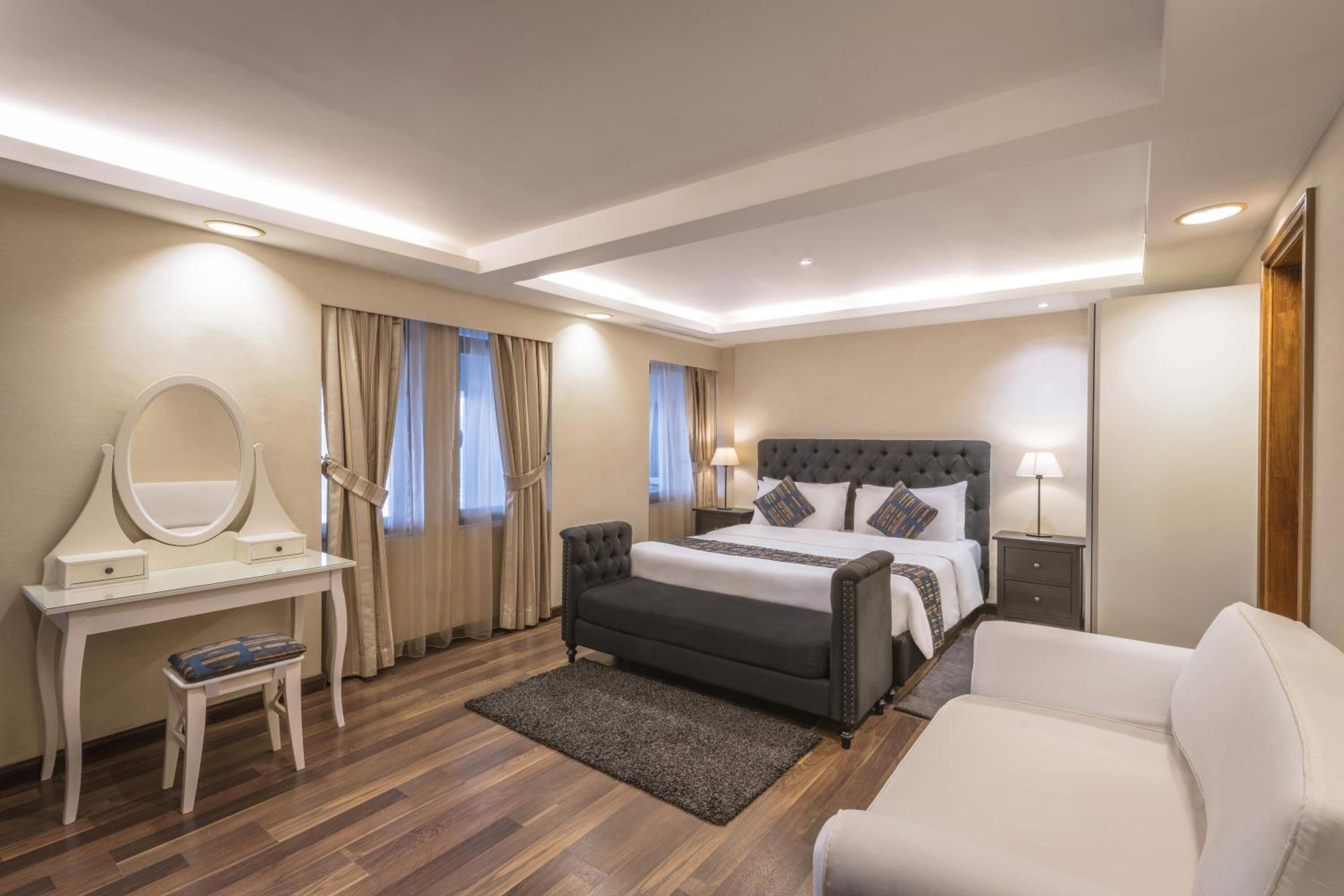 A modern bedroom with a large bed, a bedside table with a lamp, a vanity with a mirror, and a small stool.